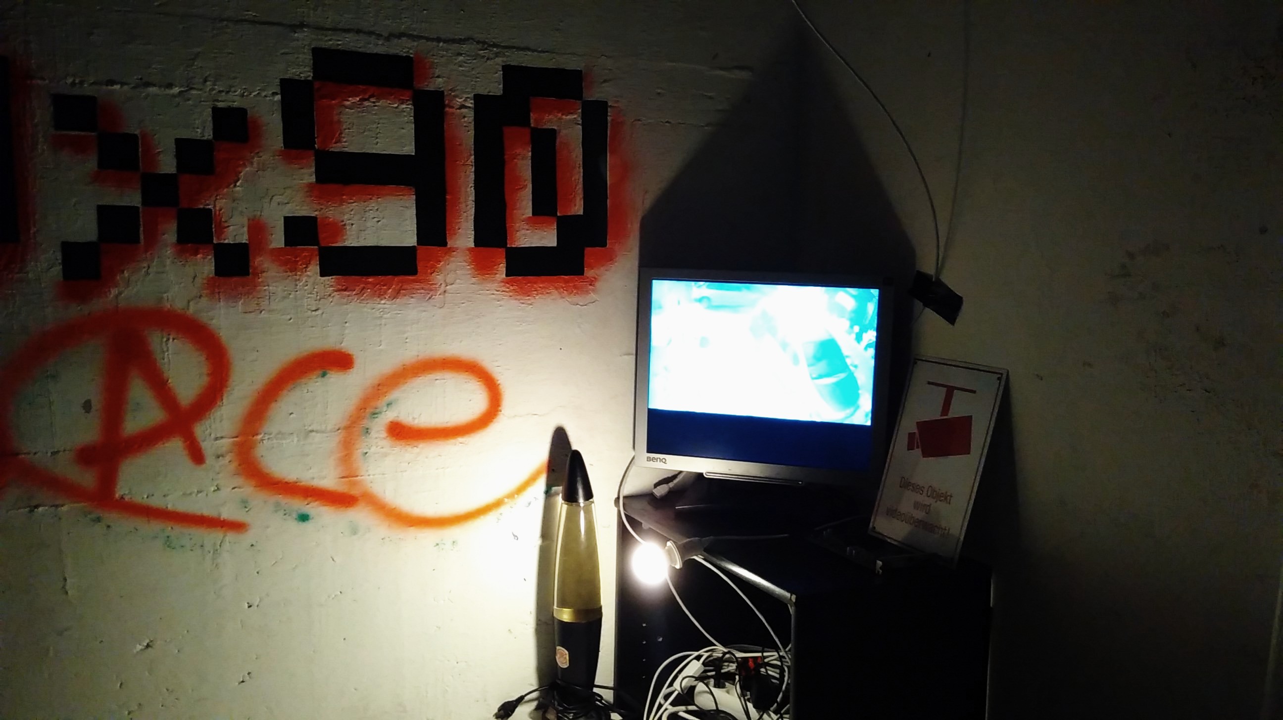 Ein leuchtender Bildschirm der neben dem 0x90.space-Schriftzug an der Wand auf einem Regal steht. Daneben sieht man eine Lavalampe und ein Schild, das vor Überwachung warnt. Das Zimmer ist abgedunkelt.