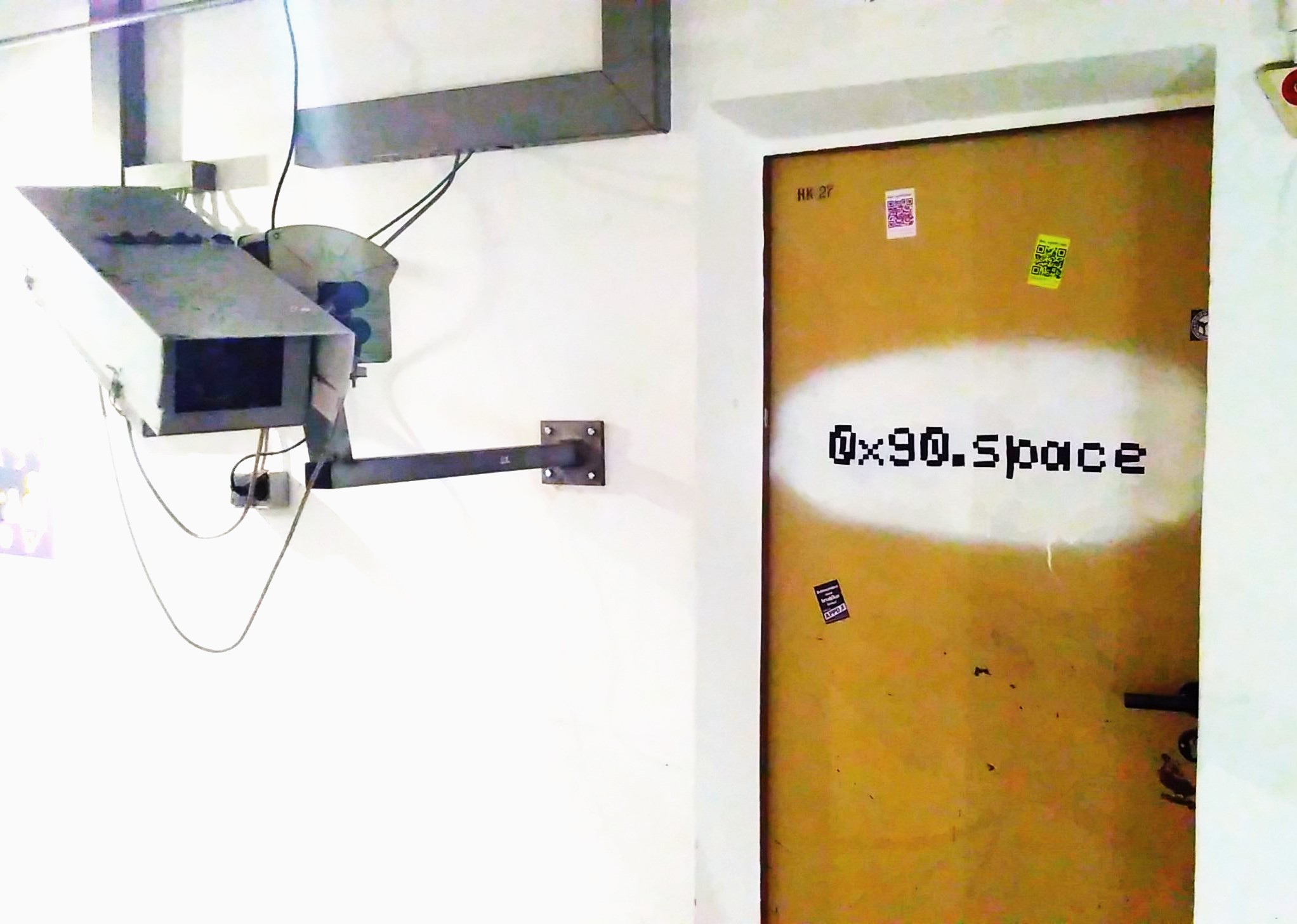 Eine Tür, an der mit schwarzer Schrift auf weißem Grund  0x90.space geschrieben ist. Links daneben ist eine überdimensionale Überwachungskamera zu sehen.